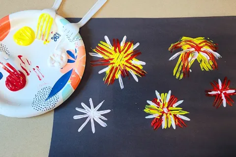 black paper with fireworks painted on it by using fork tines. a paper plate with red, yellow, and white paint and forks is next to the paper.
