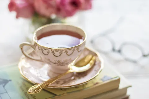 A white and gold embellished, porcelain tea cup filled with tea and saucer with a spoon sit atop a small stack of books on a marble table. 